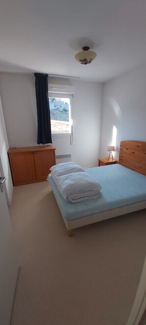 A bedroom with a bed, a wooden wardrobe, a nightstand with a lamp, a window with curtains, and a ceiling fan.