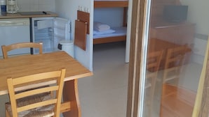 Een kleine kamer met een houten tafel, stoelen, een stapelbed, een kitchenette met koffiezetapparaat en een tv op een houten kastje.
