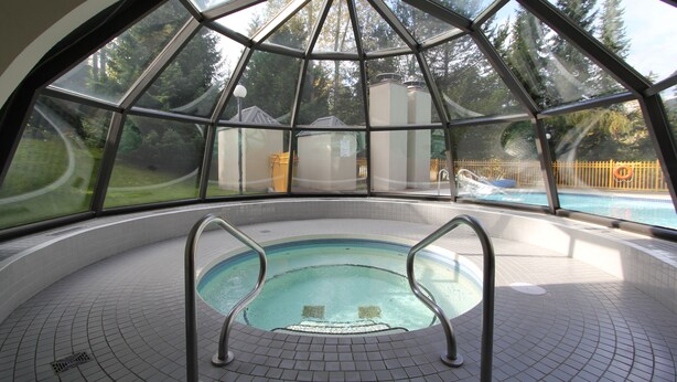 An indoor swimming pool with a glass enclosure, surrounded by a tiled floor and metal railings.
