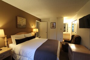 A hotel room with a bed, bedside lamps, a dresser, a chair, a TV, and a bathroom visible through an open door.