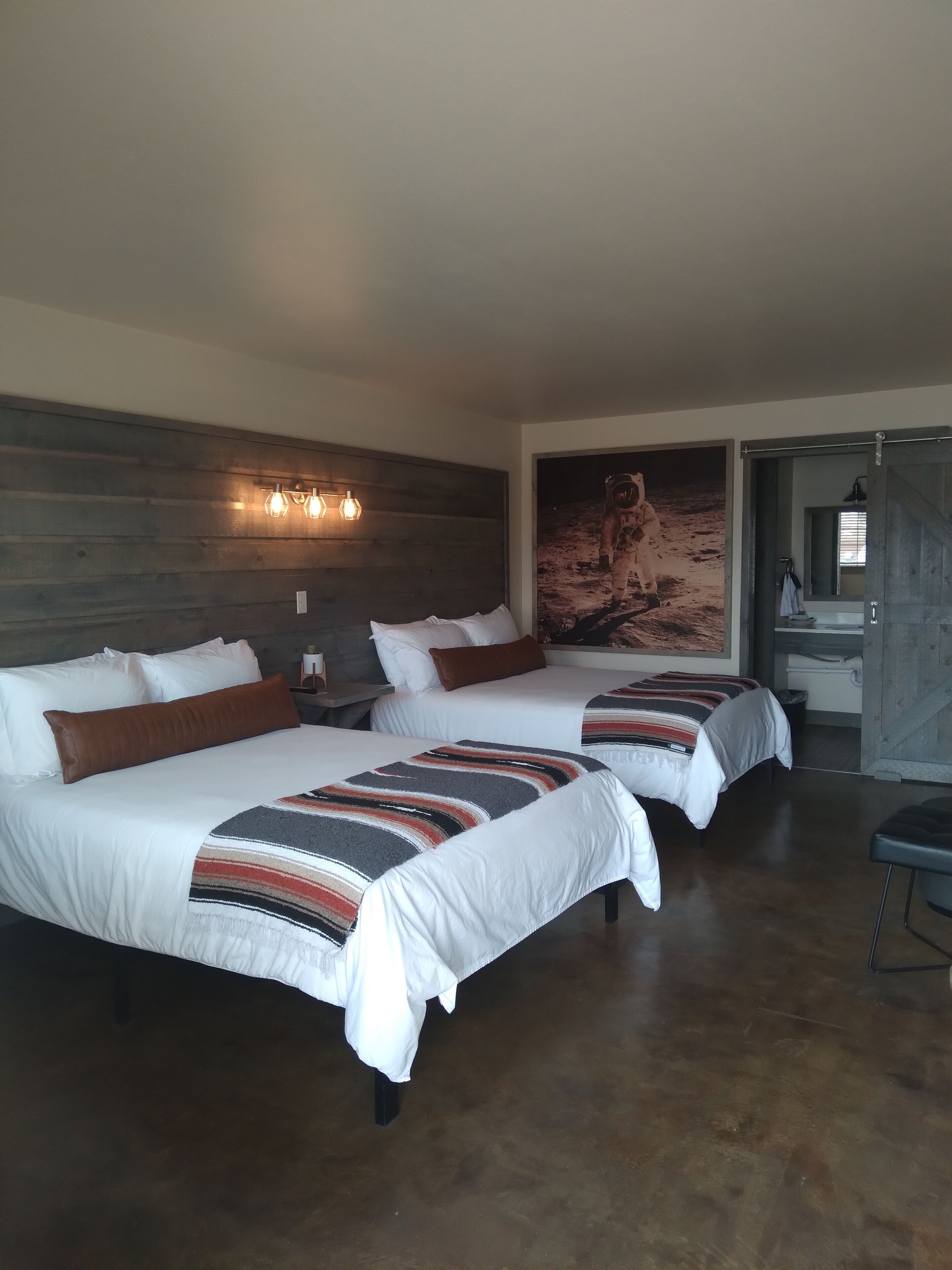 A hotel room with two beds, a wooden headboard, a large painting of an astronaut, and a bathroom visible through an open door.
