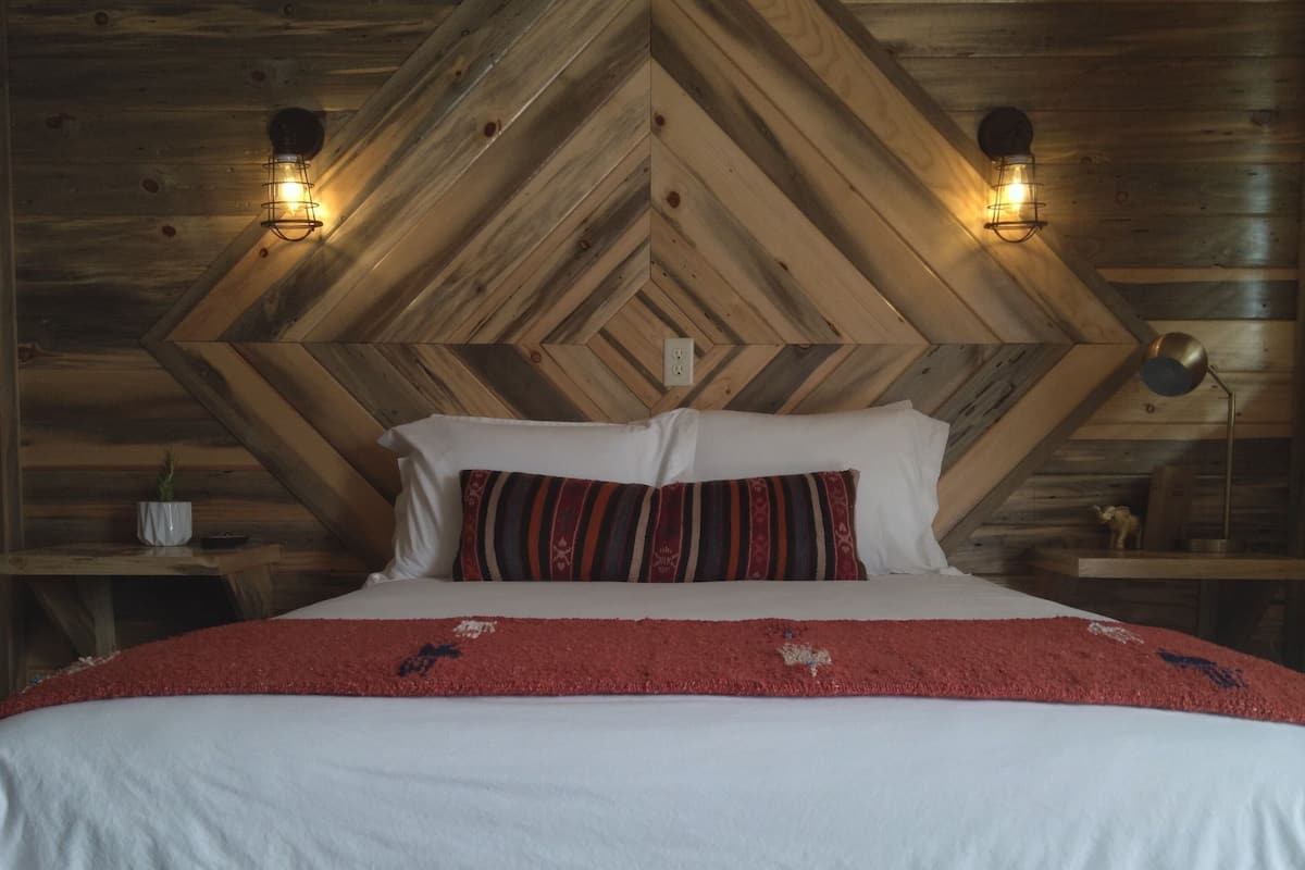 A bed with a white mattress and red blanket, flanked by two pillows, in a room with wooden walls and a chevron-patterned ceiling.