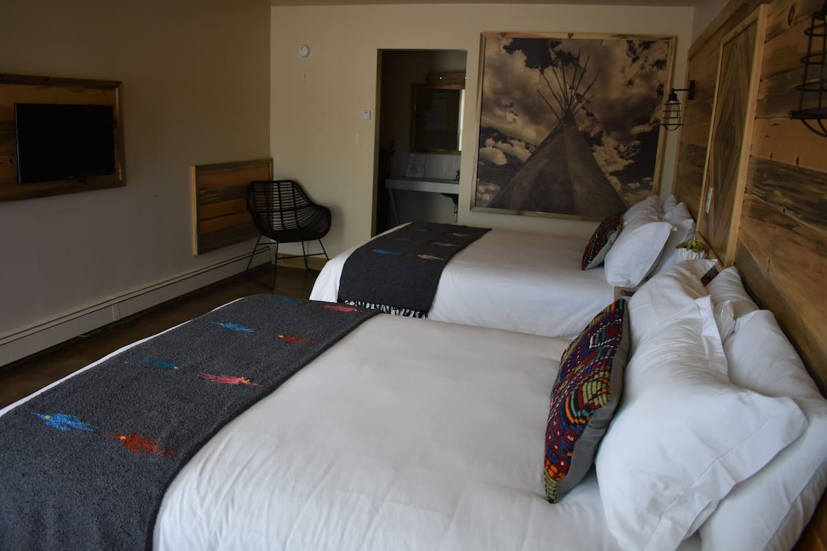A hotel room with two beds, a desk, a chair, a TV, and a painting of a tree.