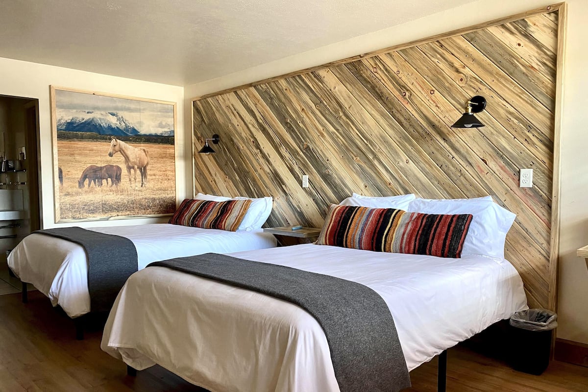 A hotel room with two beds, a wooden wall panel, a painting of horses, and a nightstand with a lamp.