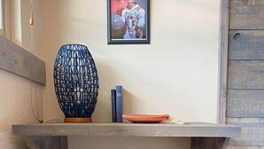Ett rum med trägolv, en metallpall, en hylla med en inramad tavla, en blå vas och två böcker.