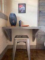 A room with a wooden floor, a metal stool, a shelf with a framed picture, a blue vase, and two books.