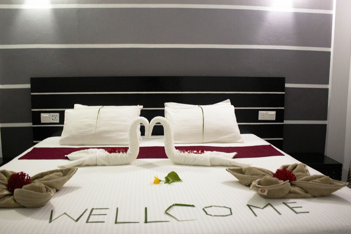 Una habitación de hotel con una cama que exhibe un letrero de toallas que dice 'WELCOME ME', dos toallas con forma de cisne y una sola flor sobre la cama.