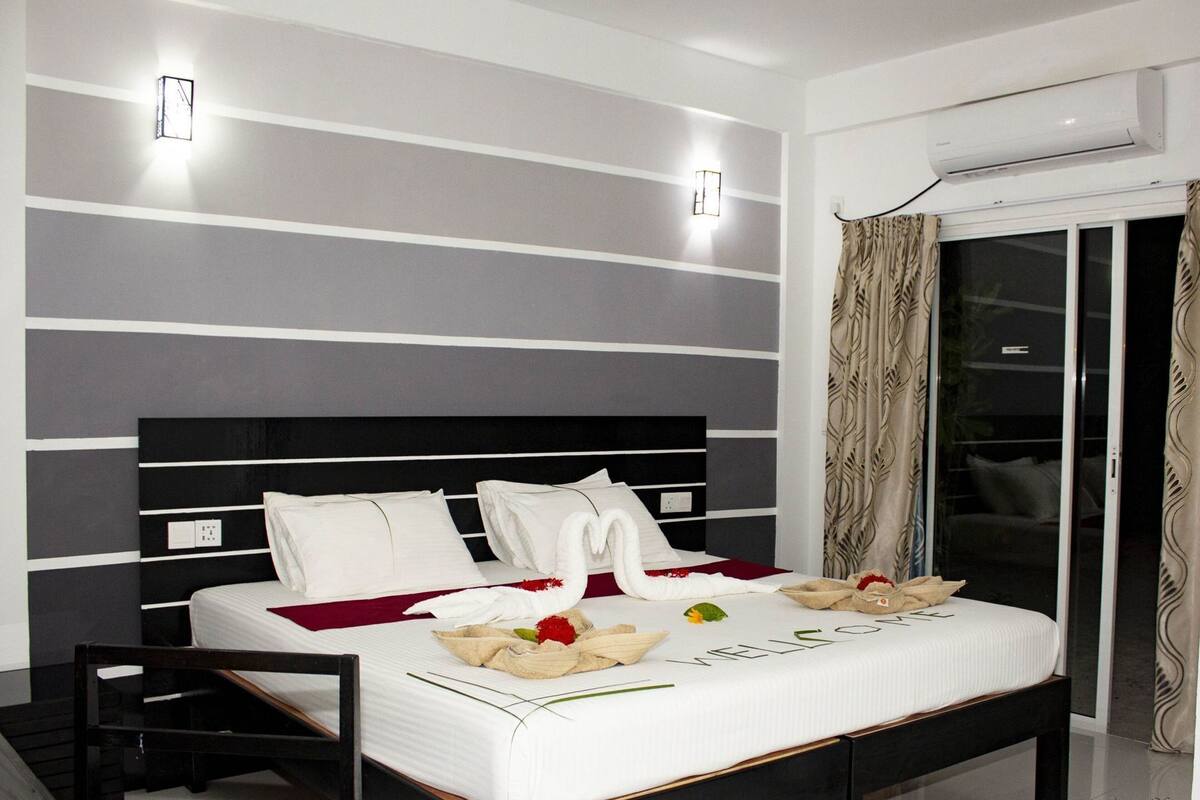 Una habitación de hotel moderna con una cama grande, una mesita de noche, aire acondicionado empotrado y una puerta de vidrio corredera con cortinas.