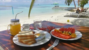 Una mesa con alimentos para el desayuno, como tostadas, huevos y fruta, con vista a una playa de aguas cristalinas y un muelle.