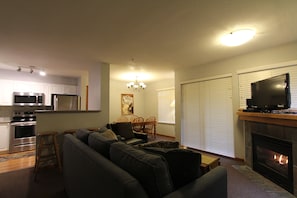 A living room with a sofa, a fireplace, a television, and a kitchen area with appliances. - Sunpath #53 | 1 Bedroom Condo with Kitchen, Fireplace, and Patio (Whistler)