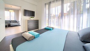 A modern hotel room with a large bed, a flat-screen TV mounted on the wall, a sofa in the adjacent room, and a view of the outdoors through sheer curtains. - Moderne Ferienwohnung in Malinska mit Pool (Milcetici)