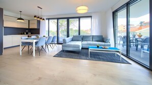 A modern living room with a grey sofa, a dining area with a table and chairs, and large windows offering a view of the outside. - Moderne Ferienwohnung in Malinska mit Pool (Milcetici)