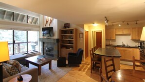 Een ruime woonkamer met een open haard, een eettafel en een keuken met houten kastjes.