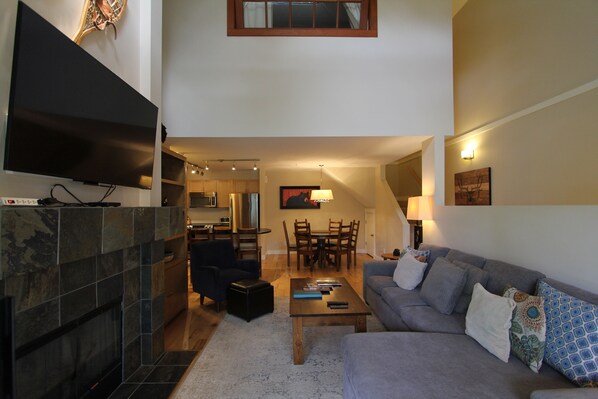 A modern living room with a flat-screen TV, a fireplace, a dining area with a table and chairs, and a gray sofa with patterned cushions.