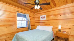 Quarto com painéis de madeira, uma cama, janela com cortinas, ventilador de teto e um quadro na parede.
