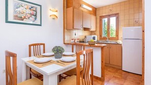 A compact kitchen with wooden cabinets, a white refrigerator, and a dining area with a table set for two. - Arena 2 bedroom villa, Cala Blanca (Ciutadella de Menorca)