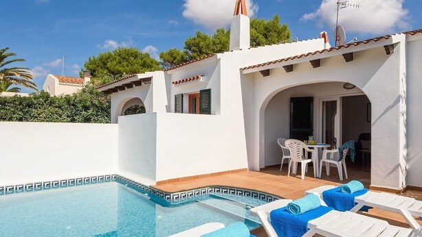 A swimming pool with lounge chairs and a table, surrounded by a white building with a tiled roof and a chimney. - Arena 2 bedroom villa, Cala Blanca (Ciutadella de Menorca)