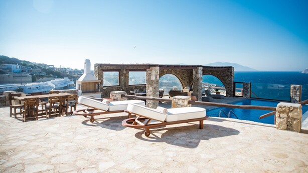 A rooftop lounge with stone flooring, lounge chairs, and a pool overlooking a coastal cityscape.