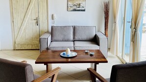 A modern living room with a wooden table, grey sofa, and two armchairs. A wooden door and a painting are visible on the walls.