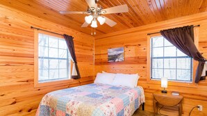 Une chambre dans un chalet en bois, avec un lit, deux fenêtres avec des rideaux, un ventilateur de plafond et une petite table avec une lampe.
