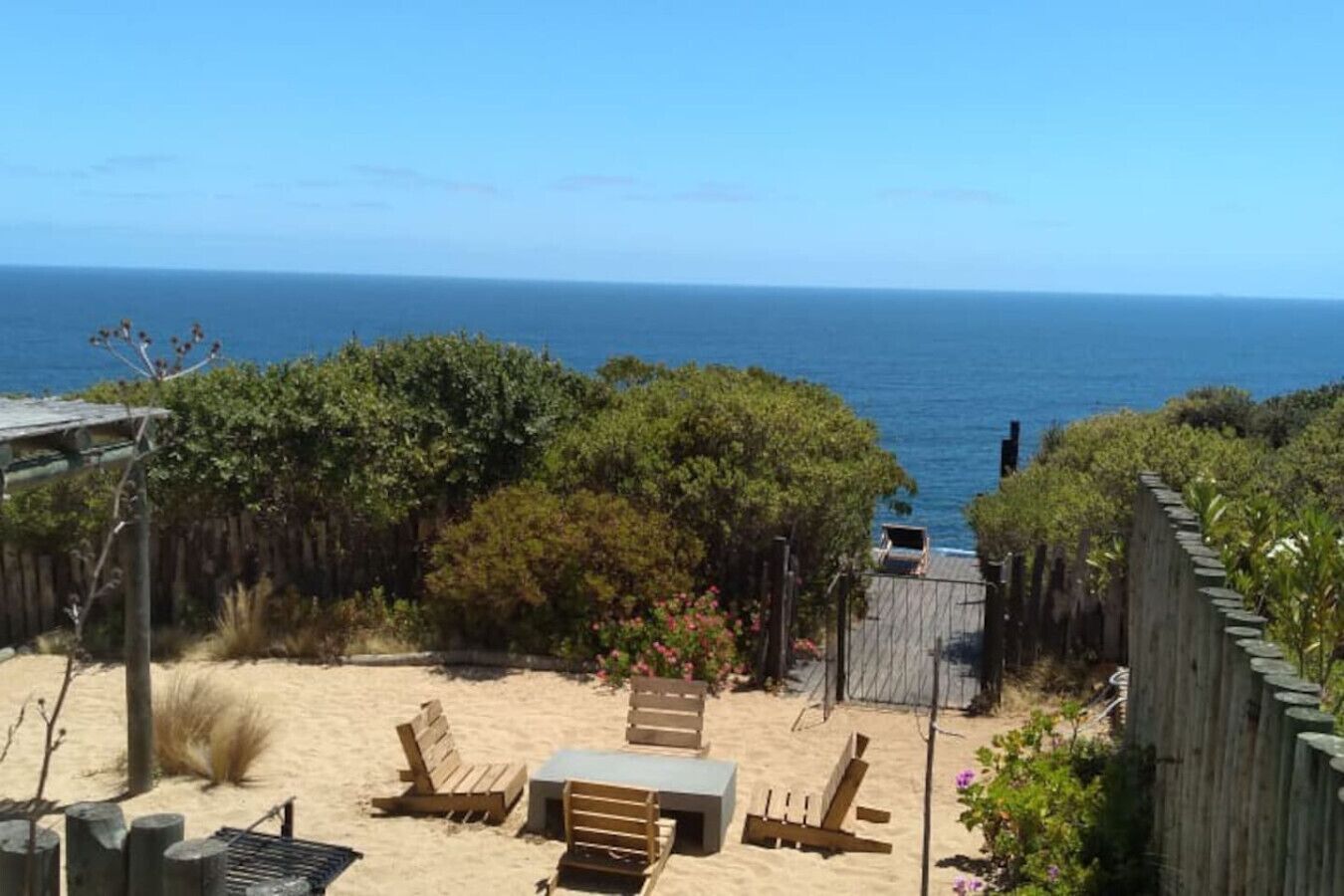 A beach with wooden chairs and a table, a wooden fence, and a view of the ocean.