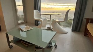 Une chambre moderne avec une table en verre, deux chaises blanches et une vue sur l’océan au coucher du soleil.