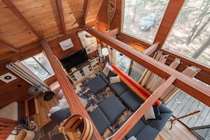 A cabin interior with a loft bed, a sofa area, a dining table, and a large window overlooking a forest. - Chill, Play & Enjoy Nature & Memories (Swanton)