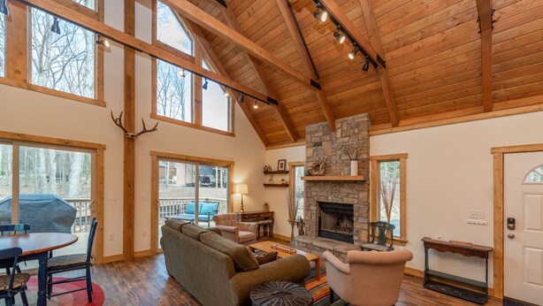 A spacious living room with a stone fireplace, wooden beams, and large windows. - Cozy Mountain Retreat w/ Hot Tub & Fire Pit (Swanton)