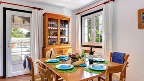 A dining area with a wooden table set for four, a cabinet with glassware, and a view through a glass door. - Ca'n Toni 3 bedroom villa, Cala Galdana (Cala Galdana)