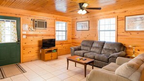 Een gezellig interieur van een chalet met houten plafond, een plafondventilator, een televisie, een bank, een bijzettafeltje en ingelijste foto's aan de wanden.