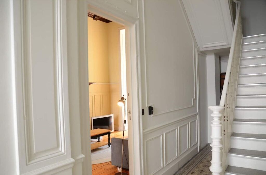 A hallway with a staircase, a door leading to a room with a television and a lamp, and a patterned floor.