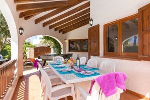 Un área de comedor al aire libre con una pérgola de madera, sillas blancas y una mesa preparada para dos.