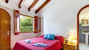 A bedroom with a large bed, a nightstand with a lamp, a window with curtains, and a bathroom visible through an archway.
