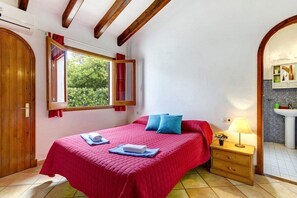 Un dormitorio con una cama grande, una mesita de noche con lámpara, una ventana con cortinas y un baño visible a través de un arco.