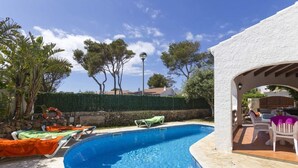 A swimming pool with lounge chairs, a pergola, and a view of trees and buildings in the background.