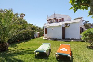 A white building with a tiled roof, surrounded by a well-maintained lawn and palm trees. There are two lounge chairs with blankets. - Gerard 4 bedroom villa, Cala Blanca (Cala Blanca)