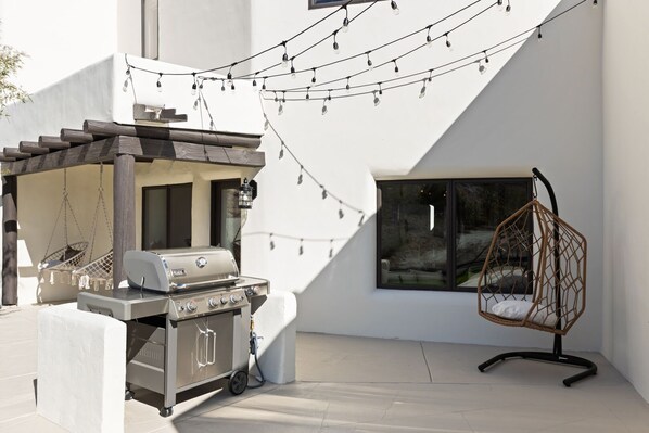 Un patio exterior moderno con una parrilla, sillas colgantes y luces de cadena.