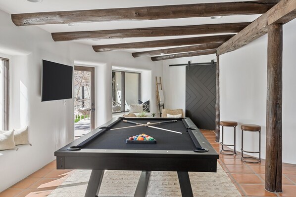 Una mesa de billar con bolas de billar, dos taburetes y un televisor montado en una sala con vigas de madera expuestas y ventanas grandes.
