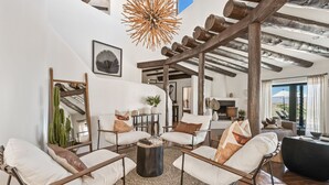 A spacious living room with a high ceiling, rustic wooden beams, a large mirror, and a view of the outdoors.