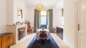 A dining area with a round table, chairs, a chandelier, a fireplace, and a view through a window.