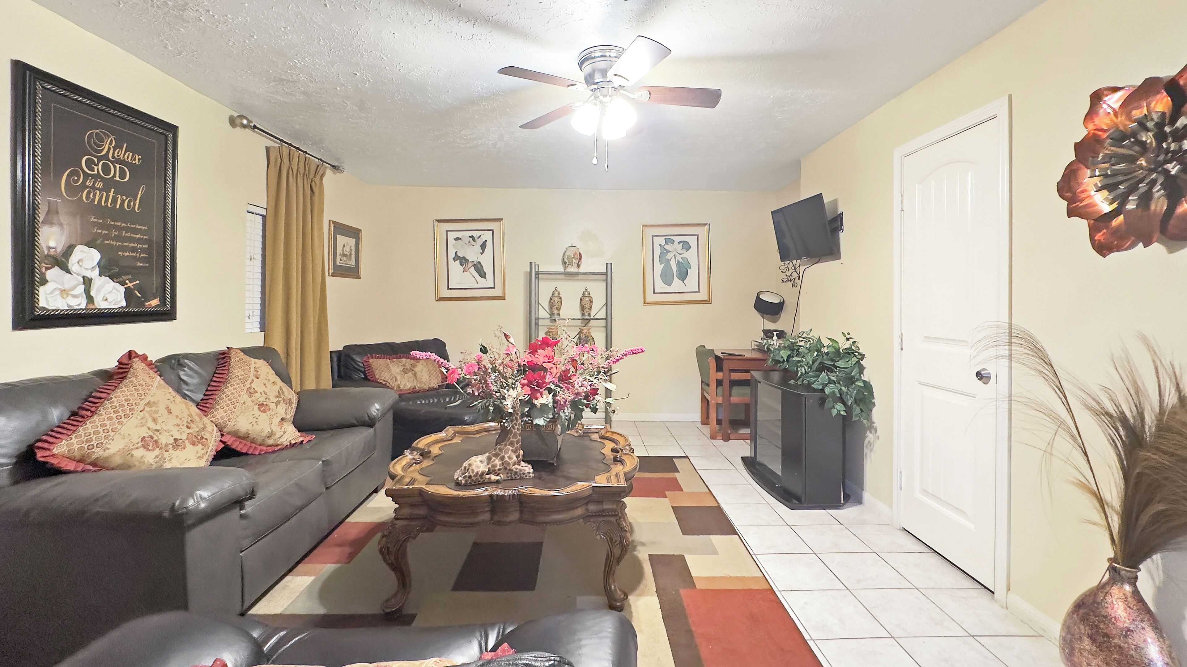 A living room with a sofa, coffee table, and a television. There is a ceiling fan, a door, and a wall with framed pictures.