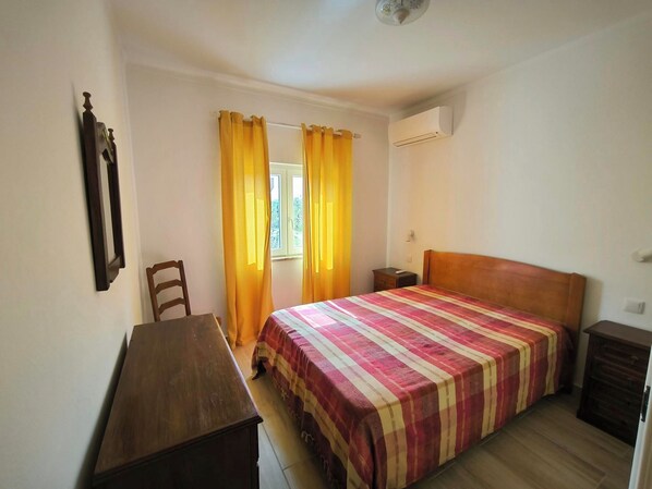 A bedroom with a bed, a wooden desk, a chair, and a small bedside table.