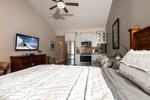 Une chambre avec un grand lit, une télévision, un ventilateur de plafond et une cuisine à l’arrière-plan.