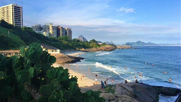 A coastal beach with people swimming and walking along the shore, surrounded by greenery and buildings in the background. - Apartamento no Coração de Copacabana (Rio de Janeiro)