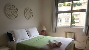 A neatly made bed with a green bedspread and two pillows, a bedside table with a lamp, and a window with a view of buildings outside. - Apartamento no Coração de Copacabana (Rio de Janeiro)