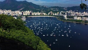 A coastal cityscape with numerous boats in a bay, a prominent mountain in the background, and dense urban development along the coastline. - Apartamento no Coração de Copacabana (Rio de Janeiro)