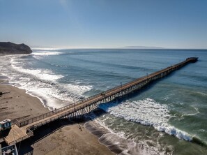 A long pier extending into the ocean, with a beach on one end and a rocky outcrop on the other.