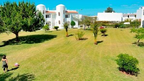 A courtyard with a large tree, a small palm tree, and a few bushes. There are white buildings with red roofs in the background. - Férias Imperdíveis , Póximo da Praia, Atrações Turisticas, bom Familias /crianç (Porches - Lagoa)