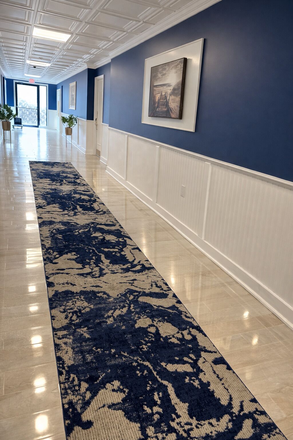 A hallway with a patterned carpet, white wainscoting, and a framed picture on the wall.