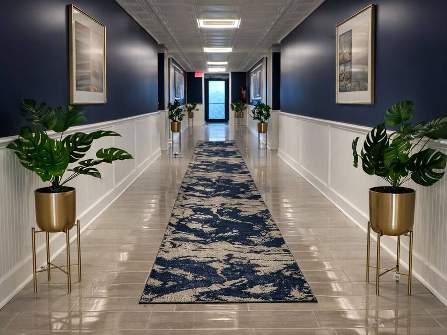 A hallway with a blue and white patterned carpet, potted plants on gold stands, framed artwork on the walls, and a white ceiling with recessed lighting.